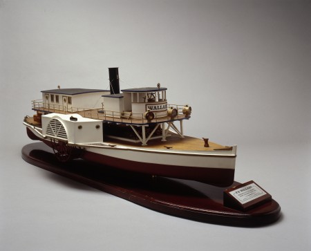 Model of a paddle steamer painted in red and white. The steamer has a wide lower deck with an upper deck with a small cabin for the helm as well as an exhaust chimney. The model is mounted on a wooden pedestal with a plaque at the bow.